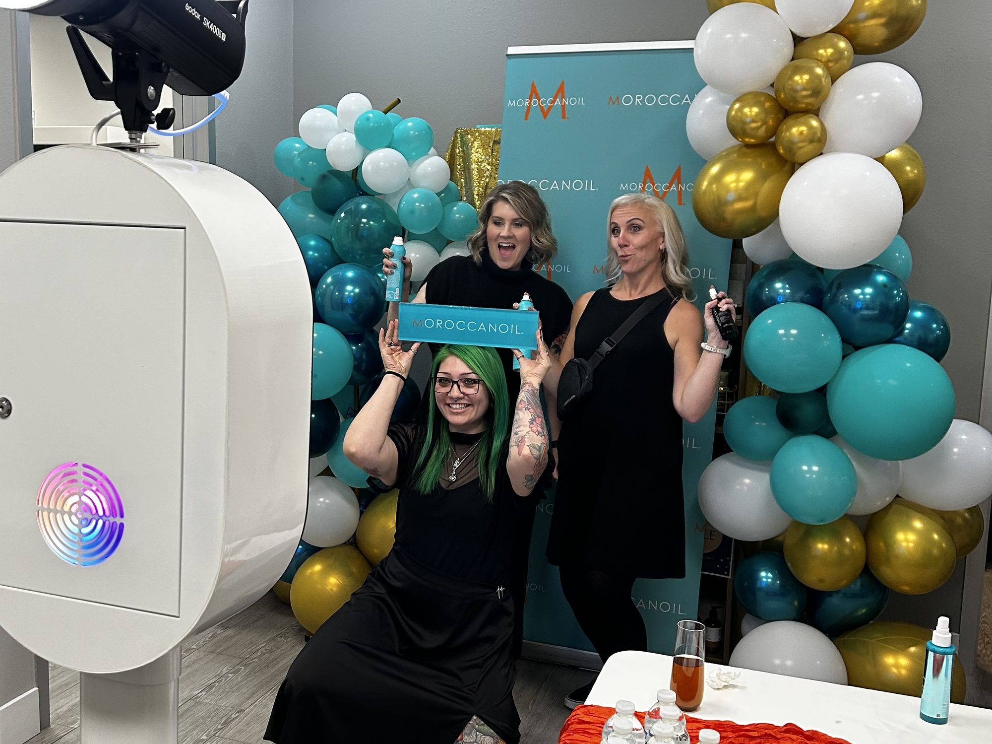 Moroccanoil branded photo booth at Sweis Inc distributor event, chrome-white booth with Moroccanoil teal-and-gold balloon arch and branded print overlays, produced by PicPop, 2025